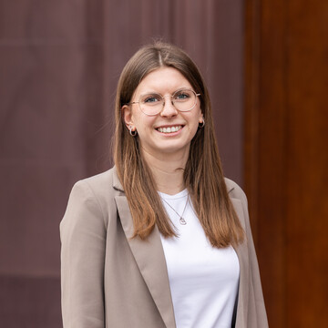 Sophie Peter hat lange braune Haare und trägt ein weißes Oberteil und einen beigen Blazer. Sie steht vor einem dunkelroten Hintergrund und lächelt.