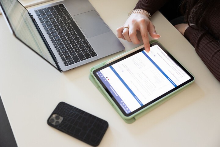 Auf einem Schreibtisch liegen Laptop und Smartphone; eine Person bedient ein Tablet.