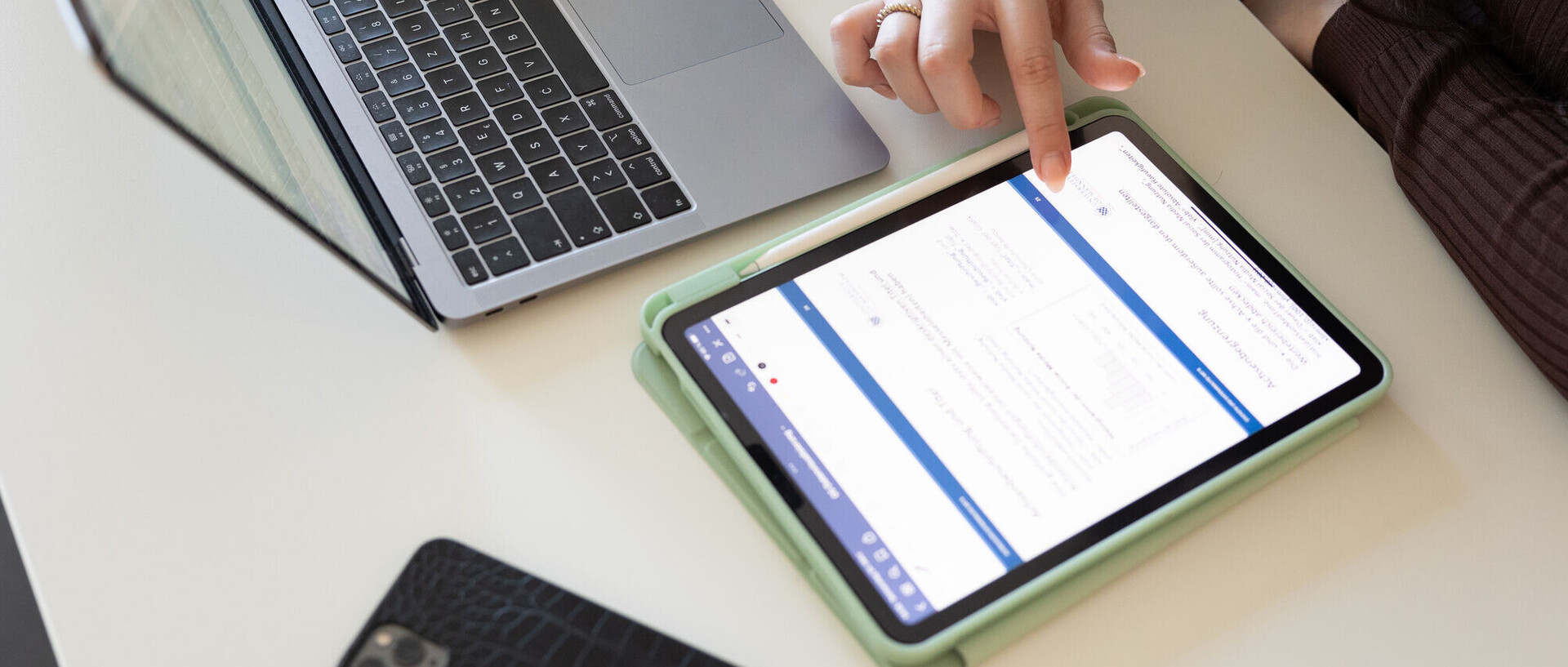 Auf einem Schreibtisch liegen Laptop und Smartphone; eine Person bedient ein Tablet.