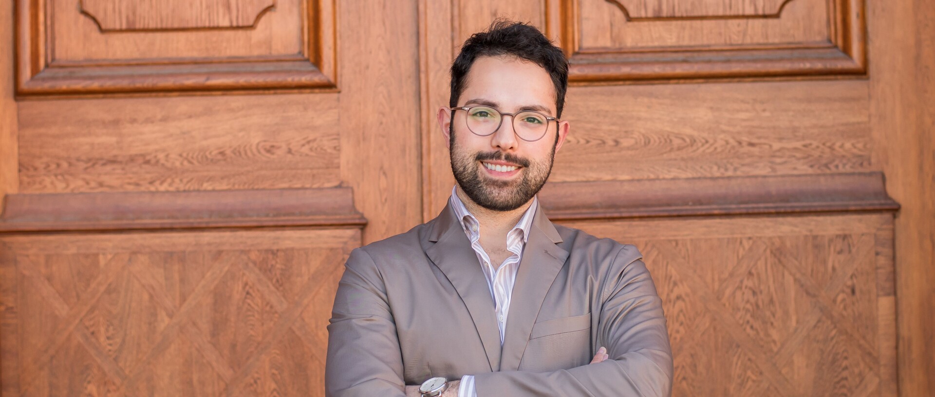 Portrait von Talha Güney. Er trägt ein beige-weiß gestreifts Hemd sowie einen beige farbenen Blazer und steht mit verschränkten Armen vor einer Holztüre.