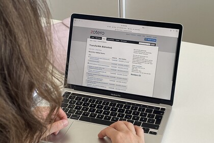 Eine Person mit schulterlangem braunem Haar sitzt an einem weißen Tisch und arbeitet an einem Laptop. Auf dem Bildschirm ist Zotero geöffnet, konkret eine Übersicht einer Bibliothek mit dem Titel ‚TransforMA Bibliothek‘. Im Hintergrund steht ein abschließbarer Aktenschrank.