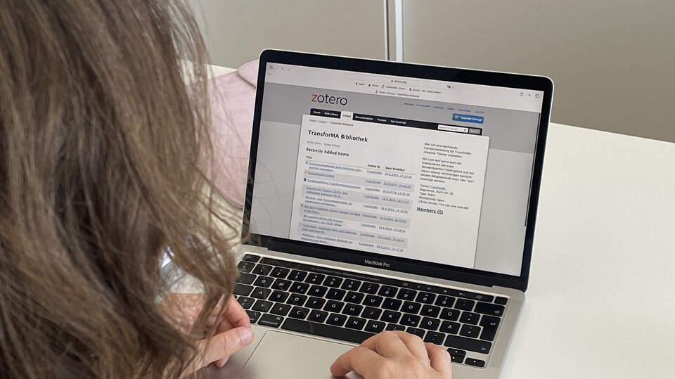 Eine Person mit schulterlangem braunem Haar sitzt an einem weißen Tisch und arbeitet an einem Laptop. Auf dem Bildschirm ist Zotero geöffnet, konkret eine Übersicht einer Bibliothek mit dem Titel ‚TransforMA Bibliothek‘. Im Hintergrund steht ein abschließbarer Aktenschrank.