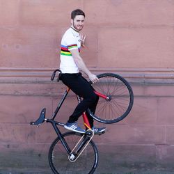 Artistic cyclist Serafin Schefold is holding the front wheel of his bicycle in the air while sitting on the handlebars. He is wearing a white shirt with colorful stripes.