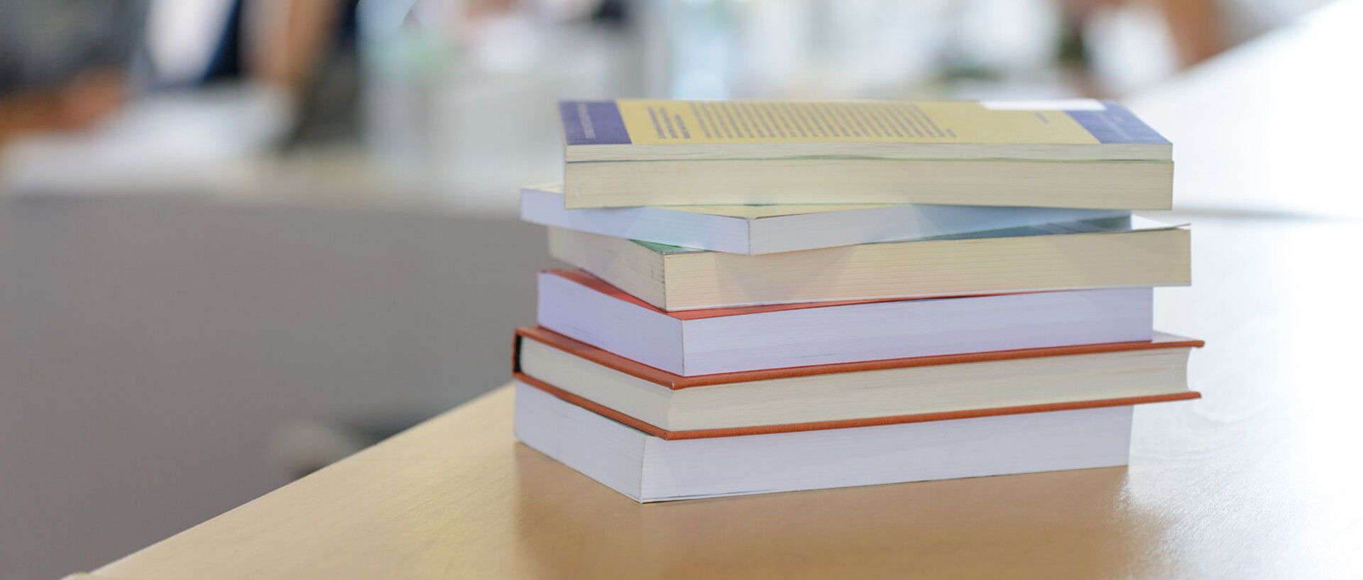 Ein Bücherstapel liegt auf einem Tisch. Im Hintergrund die Umrisse mehrerer Personen an einem Tisch.