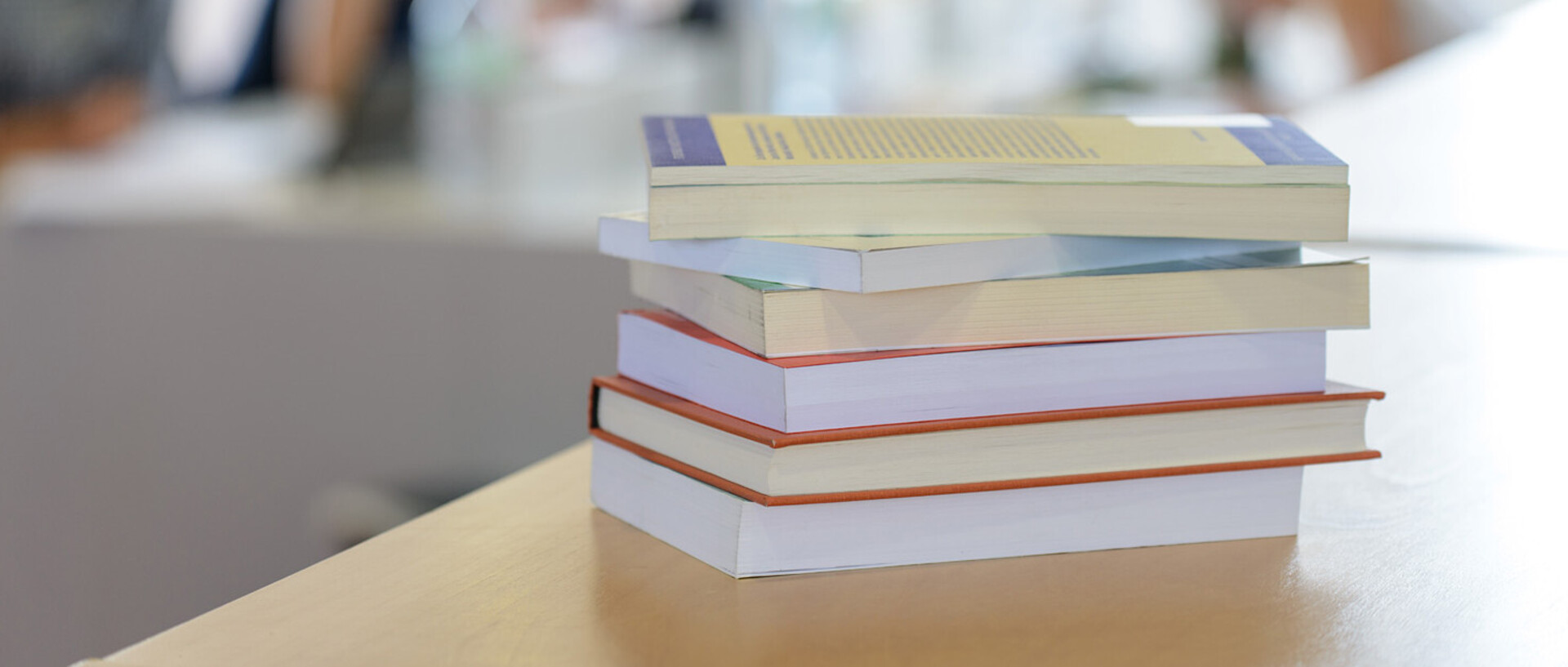Ein Bücherstapel liegt auf einem Tisch. Im Hintergrund die Umrisse mehrerer Personen an einem Tisch.
