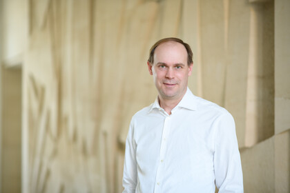 Portrait von Jannis Bischof. Er trägt ein weißes Hemd und steht vor einer beigen Wand.