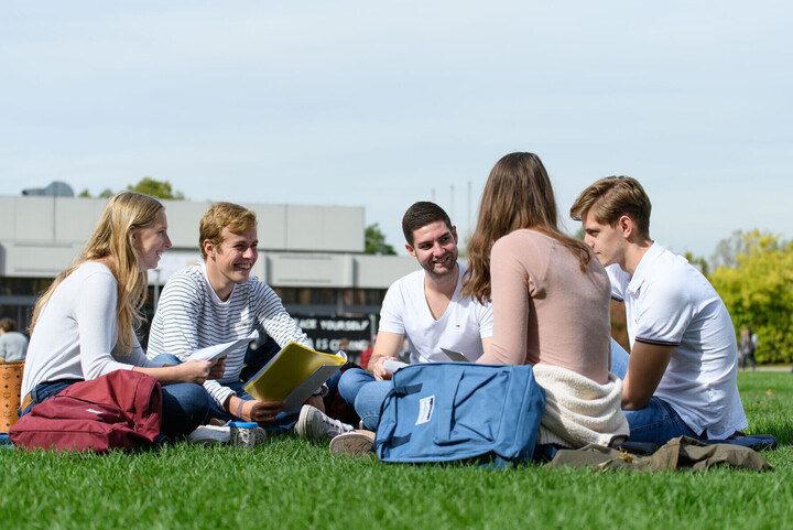 Eine Gruppe von fünf Studierenden sitzt gut gelaunt in einem Kreis auf der Mensawiese. In ihren Händen haben sie Blöcke oder Laptops, neben ihnen stehen Rucksäcke.
