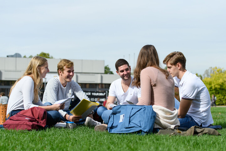 Eine Gruppe von fünf Studierenden sitzt gut gelaunt in einem Kreis auf der Mensawiese. In ihren Händen haben sie Blöcke oder Laptops, neben ihnen stehen Rucksäcke.