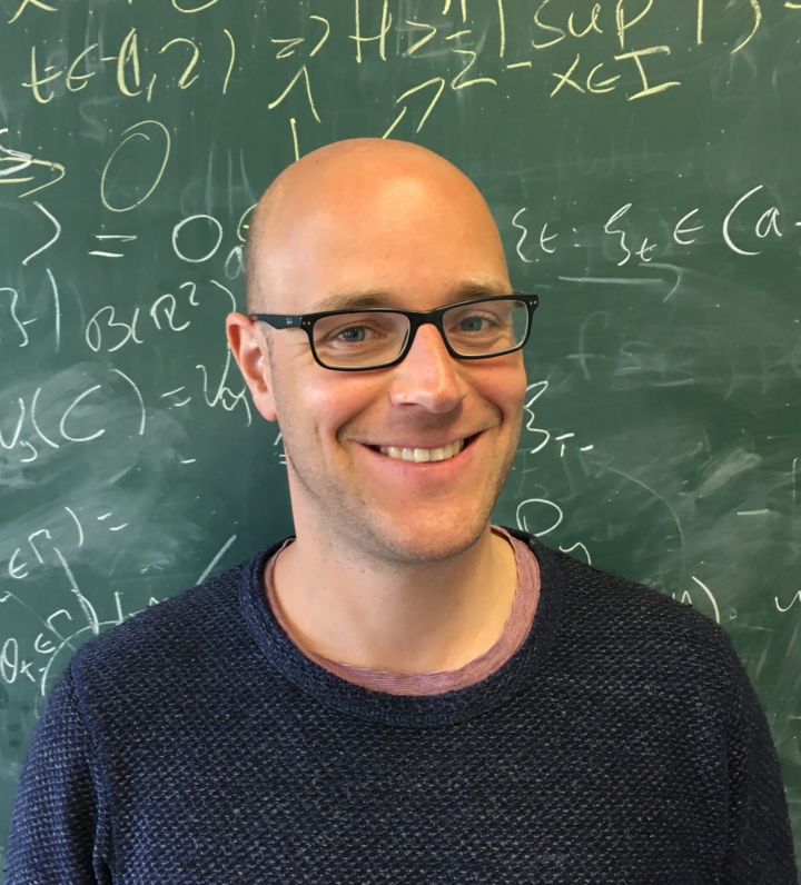 Portrait von Leif Döring. Er trägft einen grauen Pullover und steht vor einer grünen Kreidetafel.
