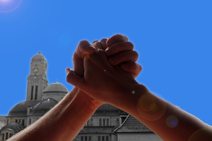 Zwei ineinander vershränkte Hände werden in die Luft gestreckt, dahinter sieht man eine Kirche.