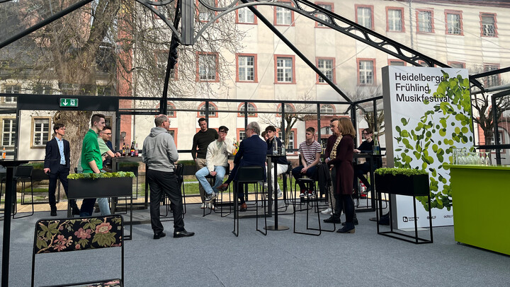 Menschen sitzen und stehen unter einem gläsernen Pavillon während einer Veranstaltung des Heidelberger Frühling Musikfestivals. Im Hintergrund sind historische Gebäude zu sehen.
