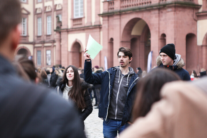 Umringt von Studierenden hält ein Campusführer auf dem Ehrenhof ein Schild mit der Aufschrift Campusführung hoch