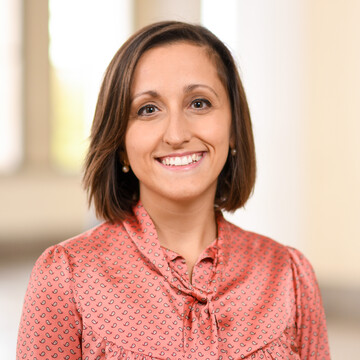 Portait of Dr. Alessandra Alloca. She is wearing a pink blouse with small white patterns on it and is standing in a room.