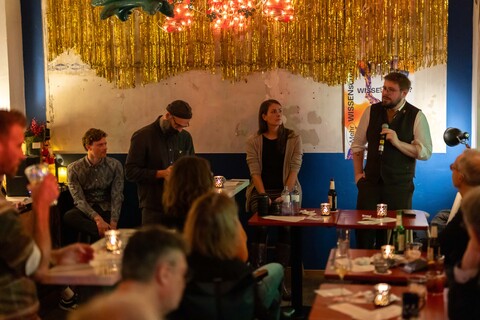 Viele Personen sitzen in einer Bar und schauen zu einer Person, die mit einer Mikrofon in der Hand spricht.