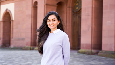 Eine lächelnde Person trägt ein weiß-blau gestreiftes Hemd unter einem lila-farbenen Pullover und steht vor dem Schloss der Uni Mannheim. Die Person heißt Areej Salah.