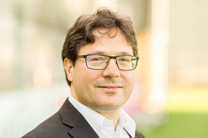 Portrait von Philipp S. Fischinger. Er trägt ein weißes Hemd sowie einen braunen Blazer und steht auf einer Rasenfläche.