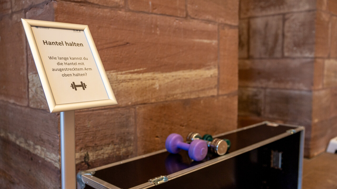 Eine große schwarze Kiste in den Katakomben der Universität, auf der drei unterschiedlich große Kurzhanteln liegen. Links daneben ein Aufsteller mit einem Schild. Auf dem Schild steht "Hantel halten. Wie lange kannst du die Hantel mit ausgestrecktem Arm oben halten?", darunter ist ein Hantel-Icon abgebildet.