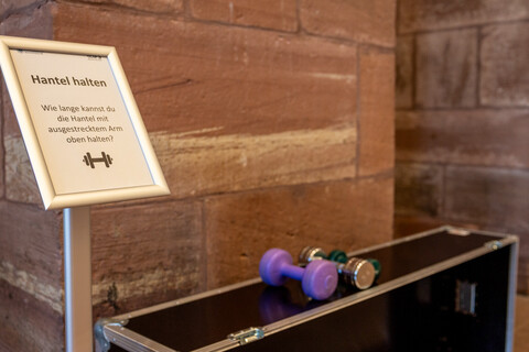 Eine große schwarze Kiste in den Katakomben der Universität, auf der drei unterschiedlich große Kurzhanteln liegen. Links daneben ein Aufsteller mit einem Schild. Auf dem Schild steht "Hantel halten. Wie lange kannst du die Hantel mit ausgestrecktem Arm oben halten?", darunter ist ein Hantel-Icon abgebildet.