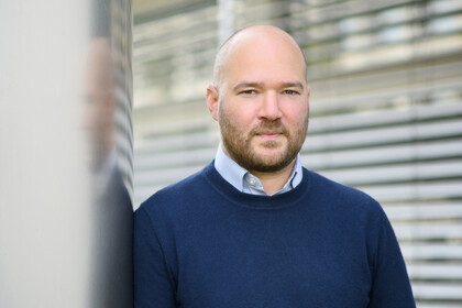 Portrait von Richard Traunmüller.Er trägt einen dunkelblauen Pullover über einem hellen Hemd und lehtn an einer grauen Säule.