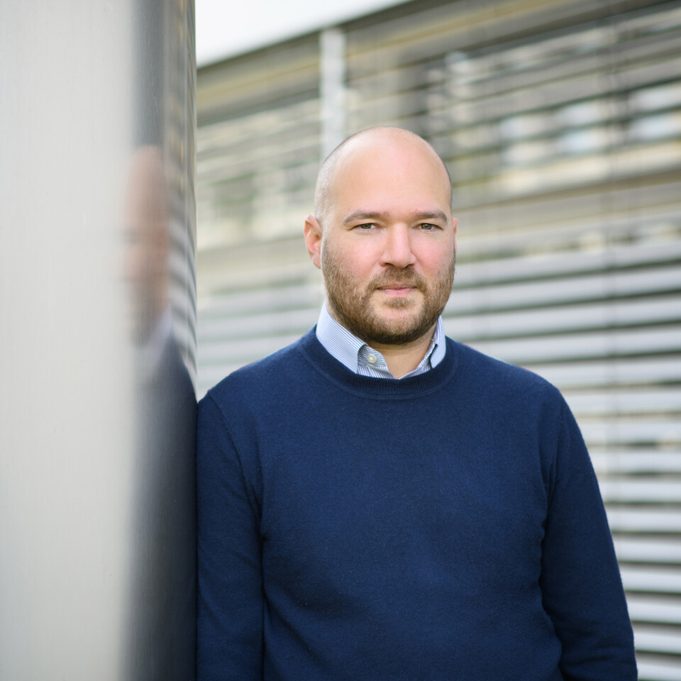 Portrait von Richard Traunmüller.Er trägt einen dunkelblauen Pullover über einem hellen Hemd und lehtn an einer grauen Säule.