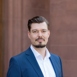 Einen Person trägt ein weißes Hemd sowie einen dunkelblauen Blazer und steht vor einer braunen Holztüre. Die Person heißt Claudius Werry.