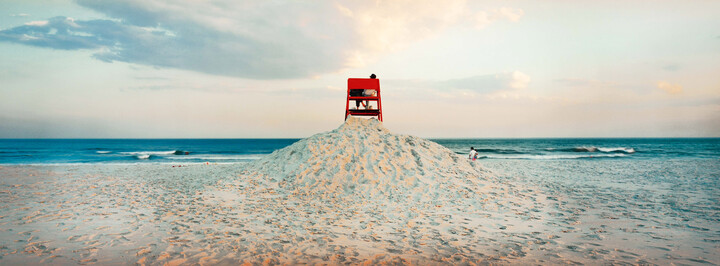 Sanstrand mit Meer im Hintergrund. Auf einem Sandhügel steht ein roter Stuhl mit zwei Personen,  Auf einem Sandhügel steht ein roter Stuhl mit zwei Personen, die von hinten zu sehen sind.