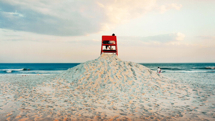 Sanstrand mit Meer im Hintergrund. Auf einem Sandhügel steht ein roter Stuhl mit zwei Personen,  Auf einem Sandhügel steht ein roter Stuhl mit zwei Personen, die von hinten zu sehen sind.