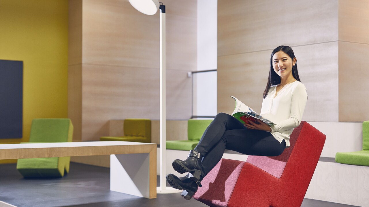 Studentin mit schwarzen langen Haaren, weißer Bluse und schwarzer Hose sitzt auf einem roten Stoffstuhl im Learning Center der Universität. Sie hat ein Magazin inder Hand und lächelt.
