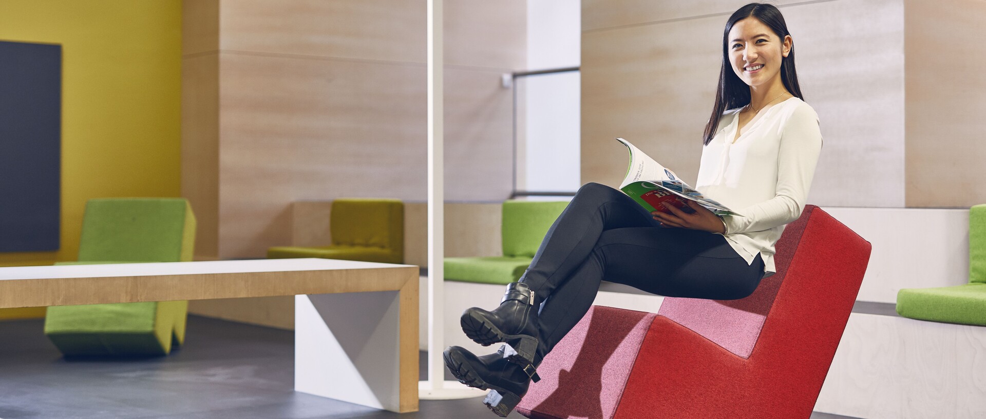 Studentin mit schwarzen langen Haaren, weißer Bluse und schwarzer Hose sitzt auf einem roten Stoffstuhl im Learning Center der Universität. Sie hat ein Magazin inder Hand und lächelt.