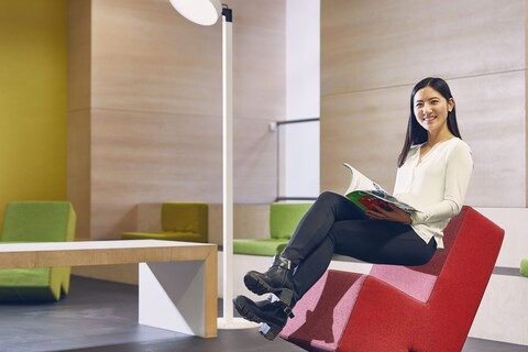 Studentin mit schwarzen langen Haaren, weißer Bluse und schwarzer Hose sitzt auf einem roten Stoffstuhl im Learning Center der Universität. Sie hat ein Magazin inder Hand und lächelt.