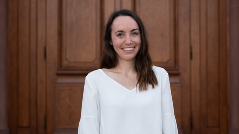 Alina Pfrang. She is wearing a white blouse and is standing in front of brown door.