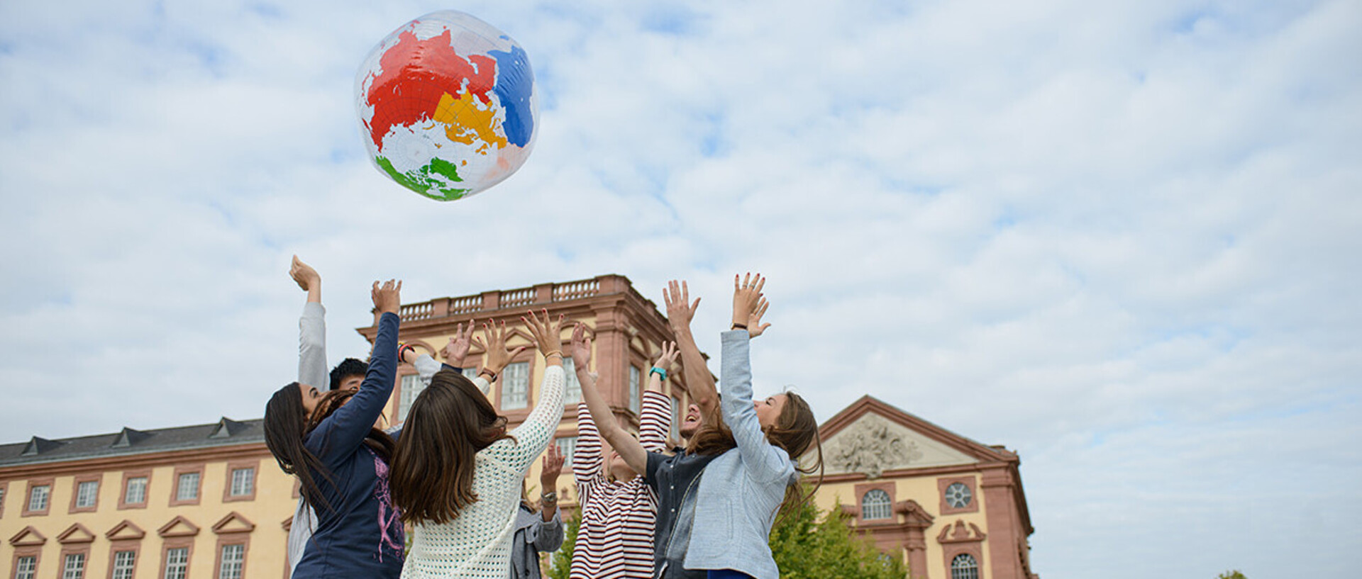 Eine Gruppe von sieben Studierenden steht in einem Kreis auf dem Ehrenhof. Sie werfen einen aufgeblasenen Globus hoch und haben die Hände in den Himmel gestreckt.