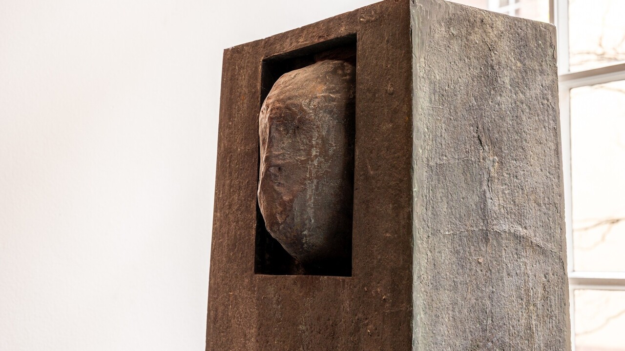 Bronzeplastik: Ein Gesicht ragt teilweise aus einem quaderförmige Block.