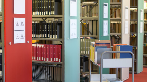 In einer Bibliothek stehen fünf Bücherregale sowie ein kleiner Rollwagen.