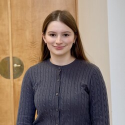 Portät der Studentin Laura Combecher. Laura Combecher trägt eine graue, vollständig zugeknöpfte Strickjacke. Ihre langen braunen Haare trägt sie offen und nach hinten über die Schulter fallend. Sie lächelt freudndlich in die Kamera.