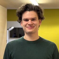 Portät des Studenten Malte Zemlin. Malte Zemlin trägt einen dunkelgrünen Pulli. Er hat braune, lockige Haare und lächelt. 