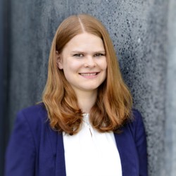 Claudia Noack. She is wearing a blue jacket over a white blouse and is leaning against a grey wall.