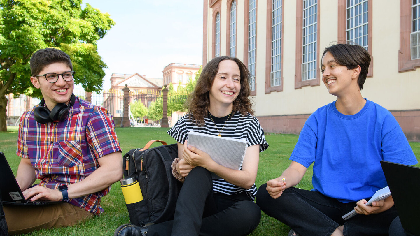 Lachende Studierende sitzen im Schneidersitz im Gras vor dem Schloss