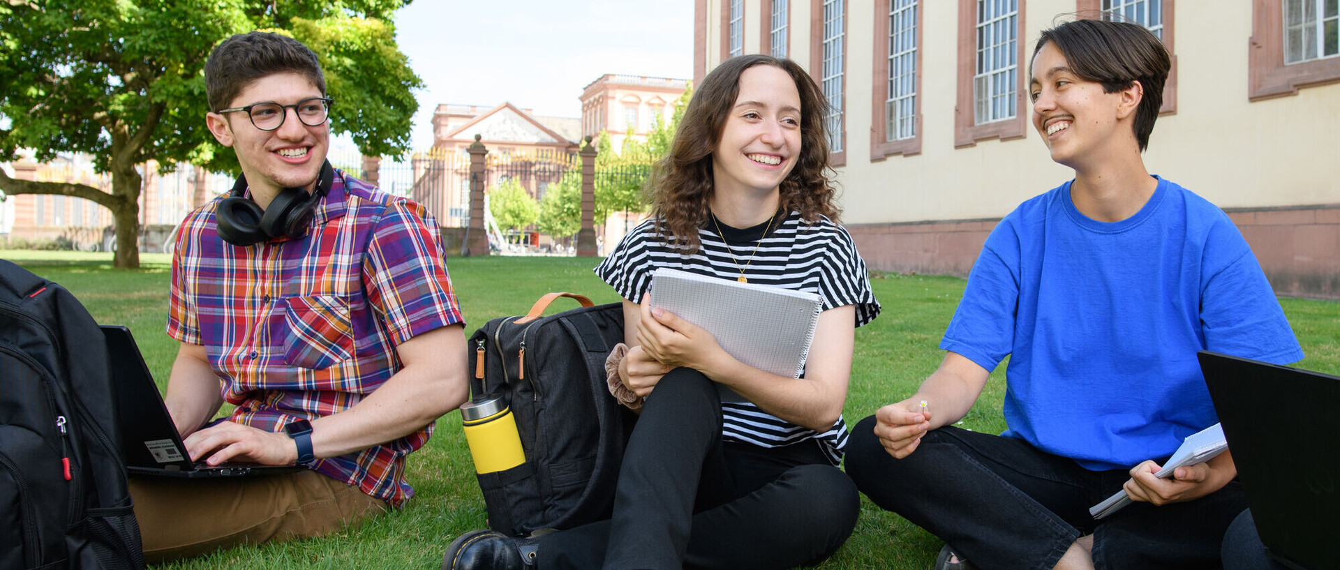 Lachende Studierende sitzen im Schneidersitz im Gras vor dem Schloss