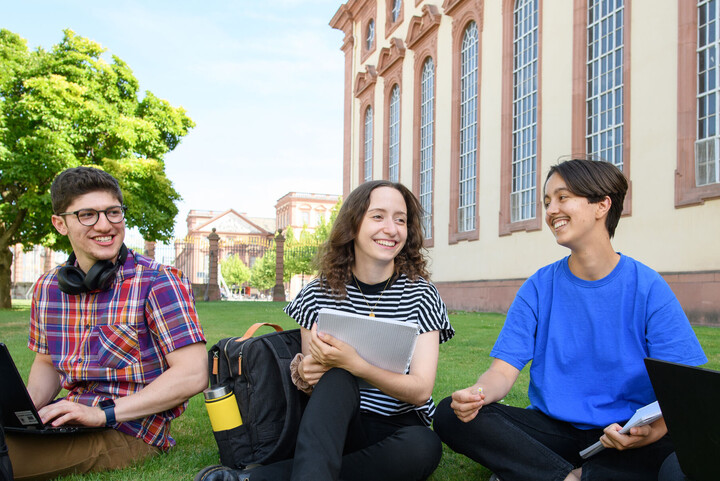 Lachende Studierende sitzen im Schneidersitz im Gras vor dem Schloss