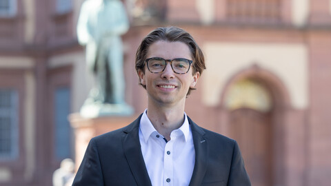 Florian Rues. He is wearing a black jacket over a white shirt and is standing on the Ehrenhof in front of one of the statues.