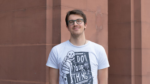 Giovanni Ballarin. He is wearing a white T-shirt and is standing in front of brown stonewall.