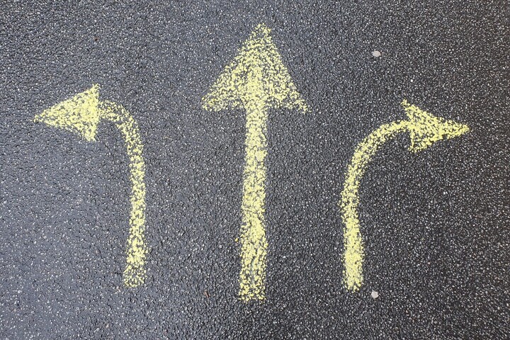Schwarzer Asphalt, darauf sind drei Pfeile mit gelber Kreide. Einer zeigt nach links, einer geradeaus, einer nach rechts.