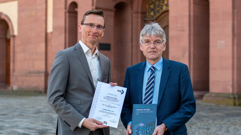 Zwei Personen in Anzug stehen nebeneinander auf dem Ehrenhof. Eine Person hält ein Lehrbuch in der Hand, die andere Person eine Urkunde.