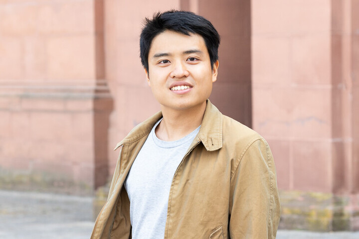 Eine lächelnde Person trägt einen grauen Pullover, eine blaue Jeans sowie eine braune Jacke und steht vor dem Schloss der Uni Mannheim. Die Person heißt Noel Chia.
