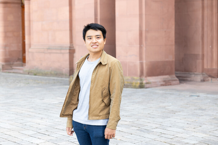 Noel Chia aus Singapur steht auf dem Ehrenhof. Er hat schwarze kurze Haare und trägt eine beige Jacke über einem grauen Shirt. Link: Noel Chia erhält DAAD-Preis