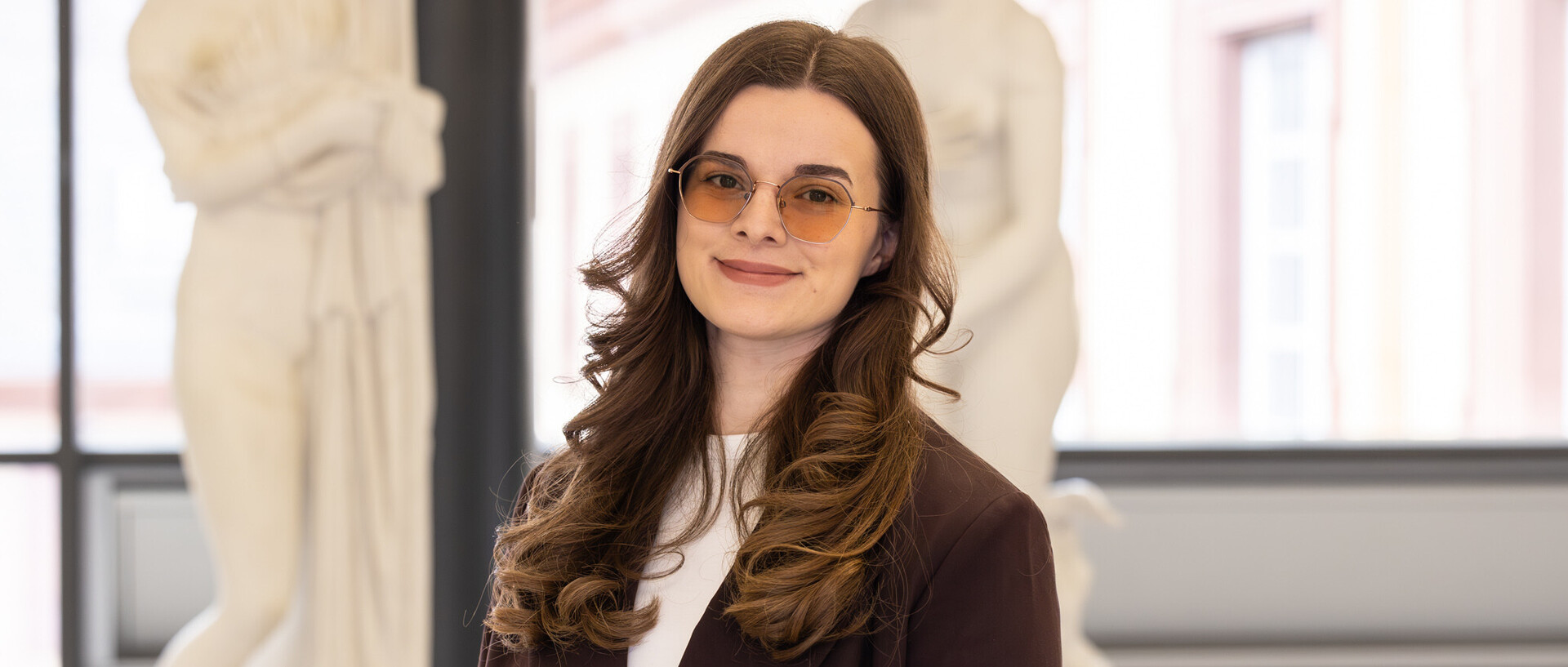 Porträt von Sara, die einen dunklen Blazer und eine getönte Brille trägt, gelockte Haare hat und vor Abbildungen antiker Statuen steht.