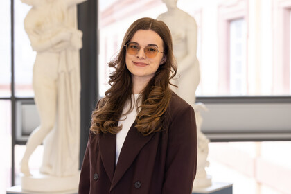 Porträt von Sara, die einen dunklen Blazer und eine getönte Brille trägt, gelockte Haare hat und vor Abbildungen antiker Statuen steht.