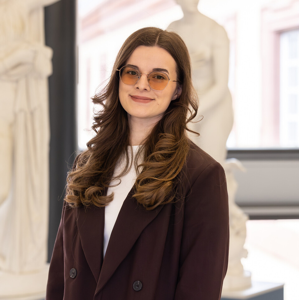 Porträt von Sara, die einen dunklen Blazer und eine getönte Brille trägt, gelockte Haare hat und vor Abbildungen antiker Statuen steht.