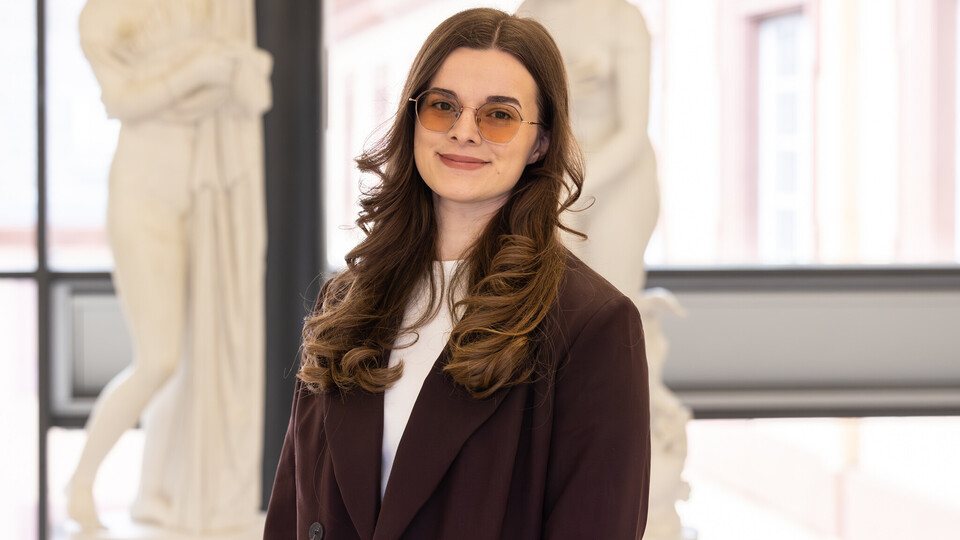 Porträt von Sara, die einen dunklen Blazer und eine getönte Brille trägt, gelockte Haare hat und vor Abbildungen antiker Statuen steht.